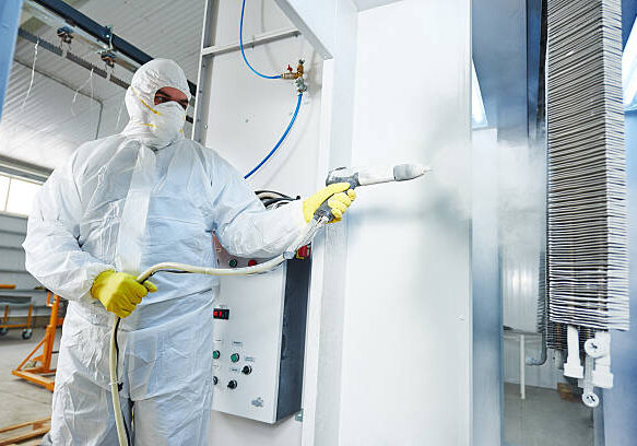 industrial metal coating. Worker man in protective suit with gas mask spraying powder to steel finished parts in painting chamber